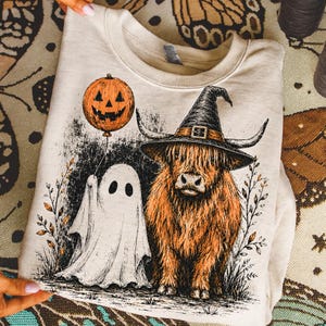 Può includere: Una felpa beige a girocollo con una stampa grafica di una mucca Highland arancione dal pelo lungo che indossa un cappello da strega nero, accanto a un fantasma bianco che tiene un palloncino a forma di zucca, circondato da fogliame nero.