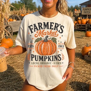 May include: A cream-colored t-shirt featuring a graphic of an orange pumpkin with green leaves. The text reads: FARMER'S Market PUMPKINS ESTD 1943 LOCAL ORGANIC - $1 EACH PICKED FRESH DAILY. The design includes small star and leaf accents.