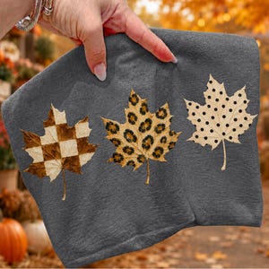 May include: A dark grey t-shirt featuring three maple leaf graphics arranged horizontally. The first leaf has a brown and cream checkerboard pattern, the second has a tan and black leopard print, and the third is cream with black polka dots.