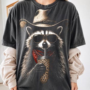 Puede incluir: Una camiseta gris carbón de manga corta con el gráfico de un mapache que lleva un sombrero de vaquero color canela y gafas de sol oscuras. El mapache tiene un pañuelo rojo estampado al cuello y sostiene un vaso con estampado de leopardo.