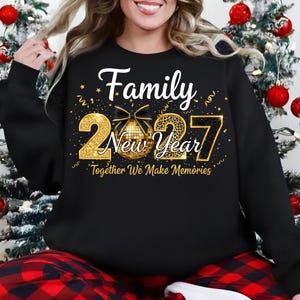 May include: A black long-sleeve sweatshirt featuring a gold graphic with the text Family New Year 2027 Together We Make Memories. The design includes a gold disco ball with a black bow, gold confetti, and the numbers 2027 in a glittery gold texture.