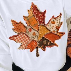 Peut inclure: Un t-shirt blanc orné d'un grand motif de feuille d'érable composé de divers empiècements en tissu aux bords effilochés. Les pièces présentent des motifs automnaux, notamment du tartan orange, des fleurs, des citrouilles et des glands.