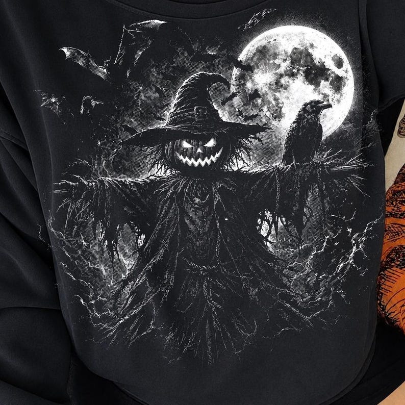 May include: A black sweatshirt featuring a detailed white graphic print of a scarecrow with a glowing jack-o'-lantern head, wearing a wide-brimmed hat. A raven sits on its arm, with bats flying against a large, bright full moon background.