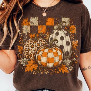 May include: A dark brown t-shirt featuring a graphic print of three pumpkins with different patterns including leopard print, orange and white checkered, and white with black polka dots, surrounded by orange autumn leaves and a checkered background.