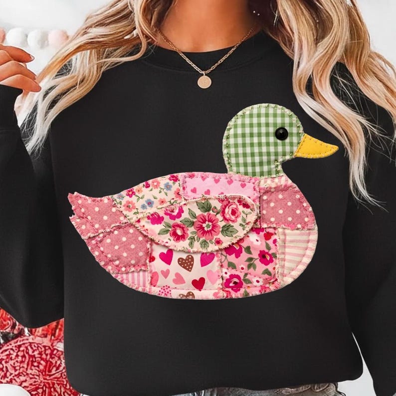 K&ouml;nnte beinhalten: Schwarzes Sweatshirt mit einem Enten-Design aus Patchwork. Die Ente hat einen gr&uuml;nen Vichy-Kopf, einen gelben Schnabel und einen K&ouml;rper aus rosa Blumen- und Polka-Dot-Stoffen. Das Design ist auf der Vorderseite des Kleidungsst&uuml;cks zentriert.