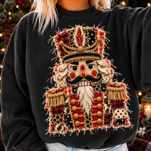 May include: A dark charcoal gray sweatshirt featuring a large, frayed-edge applique of a nutcracker. The nutcracker design incorporates various fabric patterns including red plaid, leopard print, and black polka dots with gold trim.