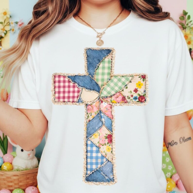 May include: White t-shirt with a patchwork cross design. The cross is made from denim, gingham, and floral fabrics, with a frayed edge. The design incorporates blue, pink, green, and red shades.