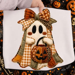 May include: A fabric ghost decoration featuring a patchwork design in shades of orange, brown, and cream. The ghost has black oval eyes, rosy cheeks, and holds a stuffed pumpkin bucket. A large bow sits atop the head, accented with floral and plaid patterns.