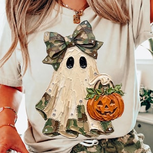 Puede incluir: Una camiseta beige con el gráfico de un fantasma con un lazo y parches de camuflaje, sosteniendo una calabaza naranja. El diseño tiene una apariencia texturizada estilo empaste con pequeños diamantes de imitación incrustados en el fantasma y el lazo.
