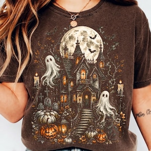 May include: A dark brown t-shirt featuring a detailed graphic print of a haunted house under a full moon. The design includes two white ghosts, several orange pumpkins, lit candles, and flying bats, creating a spooky, atmospheric scene.