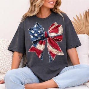 K&ouml;nnte beinhalten: Dunkelgraues T-Shirt mit einem gro&szlig;en Schleifendesign in Rot, Wei&szlig; und Blau, das an die amerikanische Flagge erinnert. Die Schleife hat eine denimartige Textur mit Sternen und Streifen. Das Shirt hat kurze &Auml;rmel und eine lockere Passform.