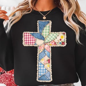 May include: Black sweatshirt with a patchwork cross design. The cross is made from denim, gingham, and floral fabrics. The cross is centrally placed on the sweatshirt, creating a unique and visually appealing design. The sweatshirt is a comfortable garment.