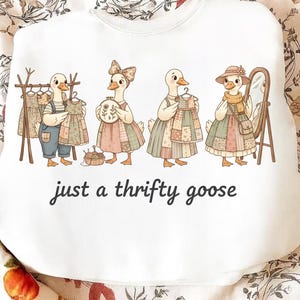 Puede incluir: Sudadera blanca con una ilustración caprichosa de cuatro gansos vestidos con vestidos de retazos, uno de ellos con un espejo. El texto "just a thrifty goose" está impreso debajo. Los gansos están representados en estilo de dibujos animados.