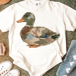 May include: Cream-colored t-shirt featuring a patchwork duck design in shades of green, brown, and blue. The duck has an orange beak and is centered on the shirt. The text "COMFORT COLORS" is printed at the top of the shirt.
