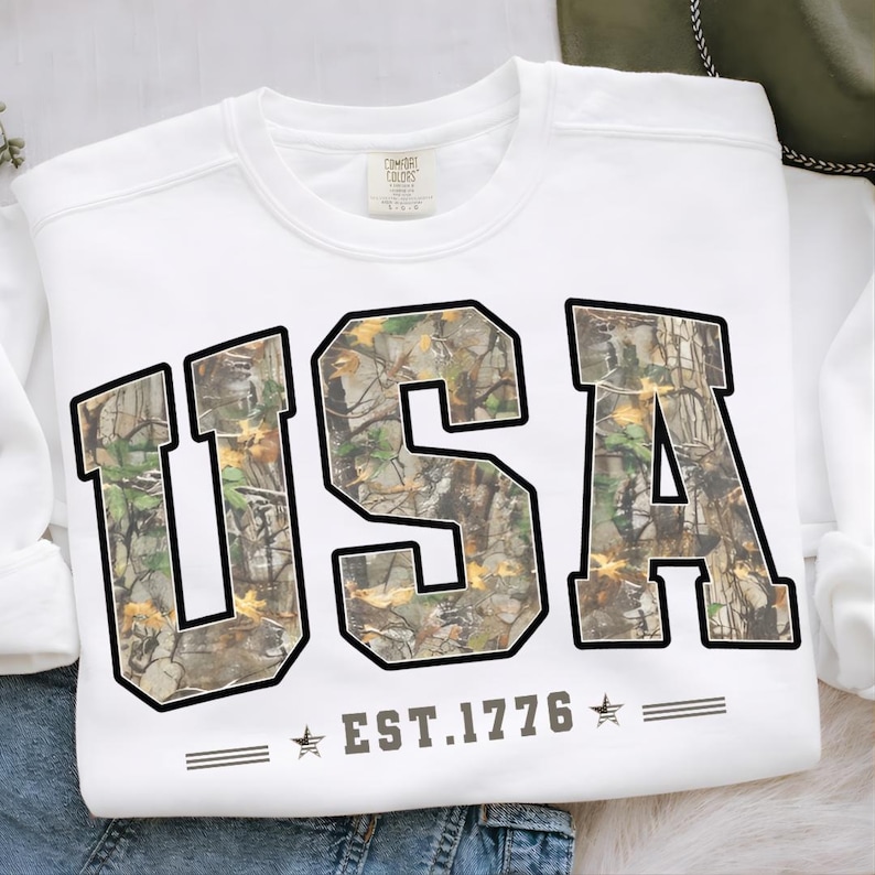 May include: White sweatshirt with the word "USA" in large, block letters filled with a camouflage pattern. Below the letters is the text "EST.1776" with stars on either side. The sweatshirt is folded on a denim surface.