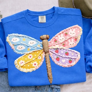May include: A blue sweatshirt with a large, colorful dragonfly applique. The dragonfly wings feature patterns of clouds, flowers, strawberries, and bunnies. The dragonfly's body is a textured brown fabric.