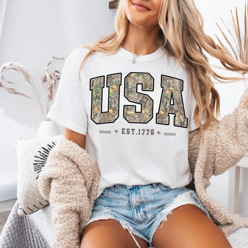 May include: White t-shirt with the word "USA" in a camouflage pattern, outlined in black. The text "EST.1776" is below the word "USA". The shirt is paired with denim shorts.