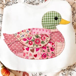 K&ouml;nnte beinhalten: Wei&szlig;es Sweatshirt mit einem aufgen&auml;hten Enten-Patchwork-Design. Die Ente hat einen gr&uuml;n-wei&szlig; karierten Kopf, einen gelben Schnabel und einen K&ouml;rper aus rosa Stoffflicken mit Blumen- und Tupfenmustern. Das Design befindet sich mittig auf dem Sweatshirt.