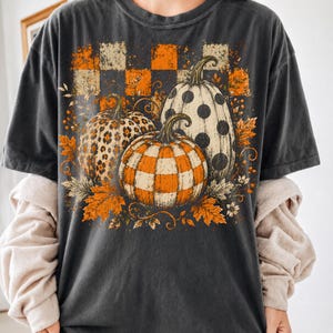 May include: A charcoal gray short-sleeve t-shirt featuring a graphic print of three pumpkins. One pumpkin has a leopard print, one has a black polka dot pattern, and one has an orange and white checkered pattern, surrounded by orange autumn leaves.