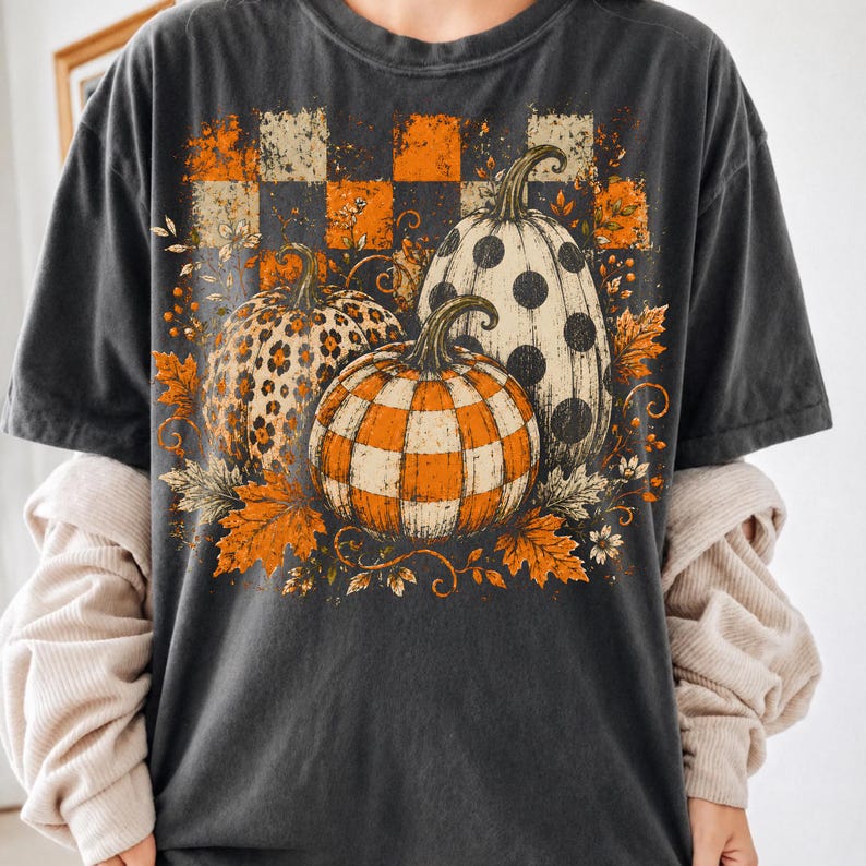 May include: A charcoal gray short-sleeve t-shirt featuring a graphic print of three pumpkins. One pumpkin has a leopard print, one has a black polka dot pattern, and one has an orange and white checkered pattern, surrounded by orange autumn leaves.