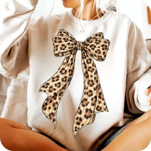 May include: Cream-colored sweatshirt featuring a large leopard print bow graphic. The bow has a detailed design with brown spots on a tan background. The sweatshirt is a casual style, suitable for everyday wear.