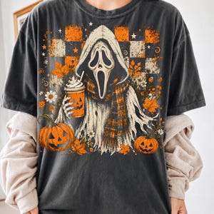 May include: A charcoal gray t-shirt featuring a graphic of a hooded ghost figure wearing a plaid scarf and holding an orange pumpkin-faced cup. The design includes orange and black checkered patterns, autumn leaves, daisies, and two jack-o'-lanterns.