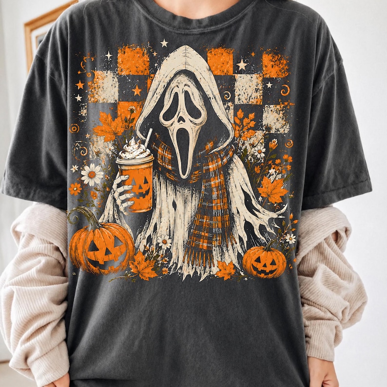 May include: A charcoal gray t-shirt featuring a graphic of a hooded ghost figure wearing a plaid scarf and holding an orange pumpkin-faced cup. The design includes orange and black checkered patterns, autumn leaves, daisies, and two jack-o'-lanterns.