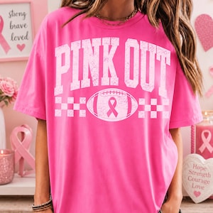 May include: A bright pink short-sleeve t-shirt featuring the white distressed text "PINK OUT" across the chest. Below the text is a white graphic of a football containing a pink awareness ribbon, flanked by two white checkered patterns.