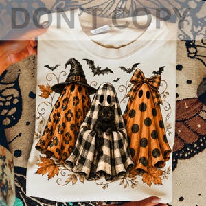 May include: A cream-colored t-shirt featuring a graphic of three ghosts. The left ghost has a leopard print pattern and a witch hat, the center ghost has a black and white buffalo plaid pattern with a black cat, and the right ghost has polka dots.