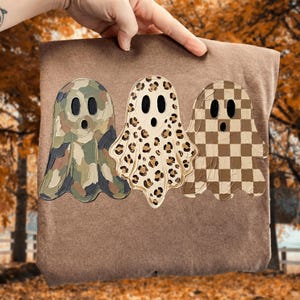 May include: A brown t-shirt featuring a graphic of three ghosts. The left ghost has a green and brown camouflage pattern, the middle ghost has a tan and brown leopard print, and the right ghost has a brown and tan checkered pattern.