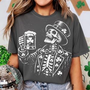 Op de afbeelding: Donkergrijs t-shirt met een witte skeletillustratie. Het skelet draagt een hoge hoed en pak, en houdt een bierpul met een klaverblad vast. Het ontwerp bevat klavers, voor St. Patrick's Day.