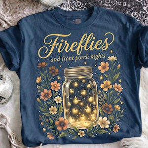Pu&ograve; includere: T-shirt blu navy con grafica di un barattolo pieno di lucciole luminose, circondato da fiori arancioni e color crema. Il testo "Fireflies and front porch nights" &egrave; arcuato sopra il barattolo in oro.