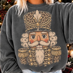 Könnte beinhalten: Ein anthrazitfarbenes Sweatshirt mit Rundhalsausschnitt und einem großen Grafikdruck eines Nussknackers. Der Nussknacker trägt eine hohe Mütze und eine Uniformjacke, die beide mit einem braun-schwarzen Leopardenmuster verziert sind.