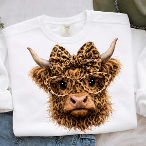 Pode incluir: Camisola branca com um close-up de uma vaca Highland com um la&ccedil;o de estampa de leopardo, &oacute;culos e pele. A vaca tem pele e chifres marrons. A camisola tem uma etiqueta Comfort Colors.