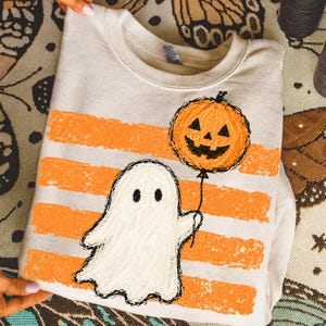 Könnte beinhalten: Ein beigefarbenes Sweatshirt mit Rundhalsausschnitt und einem Grafikdruck eines weißen Geistes, der einen orangefarbenen Kürbisballon hält. Das Design zeigt horizontale orangefarbene Pinselstrich-Streifen im Hintergrund.