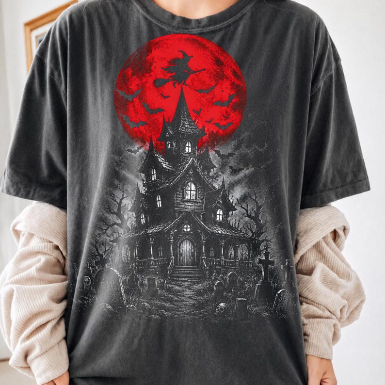 May include: A charcoal gray short-sleeve t-shirt featuring a graphic print of a dark, multi-story haunted house in a graveyard. Above the house is a large, bright red moon with a silhouette of a witch flying on a broomstick and several bats.