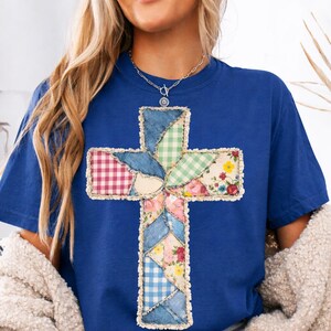 May include: A blue t-shirt with a patchwork cross design. The cross is made from denim, gingham, and floral fabrics. The edges of the cross are frayed, giving it a rustic appearance. The shirt is a solid blue colour.