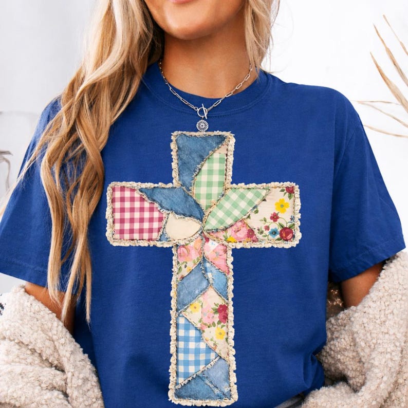 May include: A blue t-shirt with a patchwork cross design. The cross is made from denim, gingham, and floral fabrics. The edges of the cross are frayed, giving it a rustic appearance. The shirt is a solid blue colour.