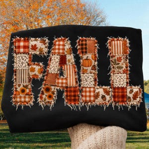 Op de afbeelding: Een zwart sweatshirt met het woord FALL in grote letters aangebracht. Elke letter is opgebouwd uit een patchwork van stoffen vierkantjes in oranje, bruine en crèmetinten, met patronen van pompoenen, zonnebloemen, bladeren en kant.