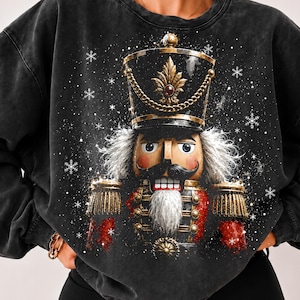 May include: A charcoal black mineral-washed sweatshirt featuring a large graphic print of a nutcracker soldier. The nutcracker wears a tall black hat with gold trim, a red uniform with gold buttons, and has white hair and a white beard.