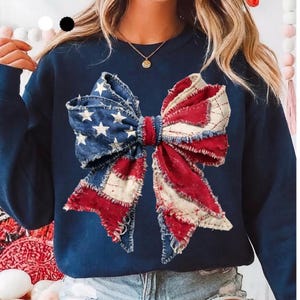 May include: Navy blue sweatshirt featuring a large bow design. The bow is crafted from fabric with a distressed, patriotic theme, incorporating red, white, and blue colors. The bow's design includes stars and stripes, evoking an American flag.