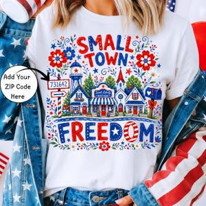 May include: A white t-shirt featuring a graphic print with the text SMALL TOWN FREEDOM. The design includes a small town scene with a water tower, a boutique, a mailbox, and a signpost with the text 731642, surrounded by red and blue floral accents.