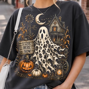 May include: A charcoal gray t-shirt featuring a graphic print of a ghost with a leopard print pattern, a wooden sign reading I LIVE HERE NOW, a jack-o'-lantern, a haunted house, a crescent moon, a windmill, and a vintage lantern in orange and black.