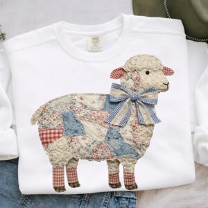 Könnte beinhalten: Weißes Sweatshirt mit einem Patchwork-Schaf-Design. Das Schaf ist mit floralen, Vichy- und Spitzenstoffen, einer blauen Vichy-Schleife und roten Vichy-Akzenten an Ohren und Beinen verziert. Das Sweatshirt hat einen Rundhalsausschnitt.