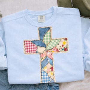 May include: A pale blue sweatshirt with a patchwork cross design. The cross is made from denim, gingham, and floral fabrics, edged with gold-colored trim. The sweatshirt has long sleeves and a crew neck.