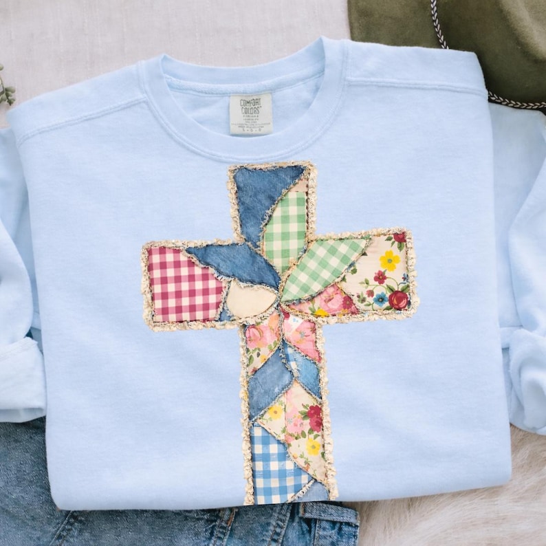 May include: A pale blue sweatshirt with a patchwork cross design. The cross is made from denim, gingham, and floral fabrics, edged with gold-colored trim. The sweatshirt has long sleeves and a crew neck.