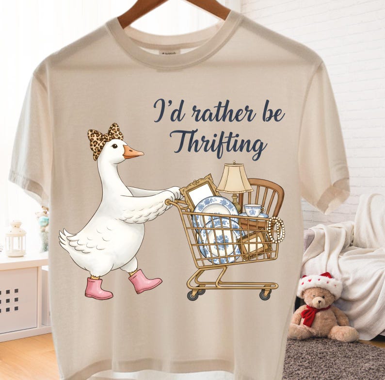 Puede incluir: Camiseta beige con un ganso de dibujos animados con un lazo de leopardo y botas rosas, empujando un carrito de compras lleno de art&iacute;culos vintage. El texto "I'd rather be Thrifting" est&aacute; impreso en la camiseta.