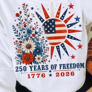 May include: A white t-shirt featuring a graphic of a sun with an American flag pattern, surrounded by red, white, and blue flowers and stars. The text reads 250 YEARS OF FREEDOM, 1776, and 2026, with a distressed, vintage-style print design.