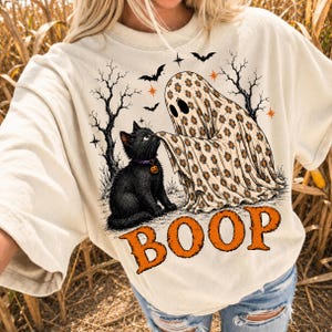 May include: A cream-colored t-shirt featuring a graphic of a black cat and a ghost covered in a floral pattern. The ghost is touching the cat's nose. The design includes bare trees, bats, stars, and the orange text BOOP at the bottom.