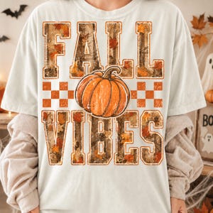 May include: A cream-colored short-sleeve t-shirt featuring the text FALL VIBES in block letters with a camouflage and autumn leaf pattern. An orange pumpkin graphic is centered between the words, accompanied by an orange and white checkerboard.